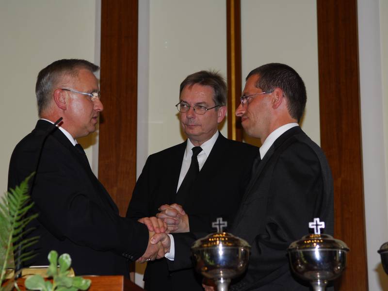 Bezirksapostel Nadolny (l.) entbindet Bezirksevangelist Kasper (r.) von seiner Vorsteheraufgabe und beauftragt damit neu den Evangelisten Lange